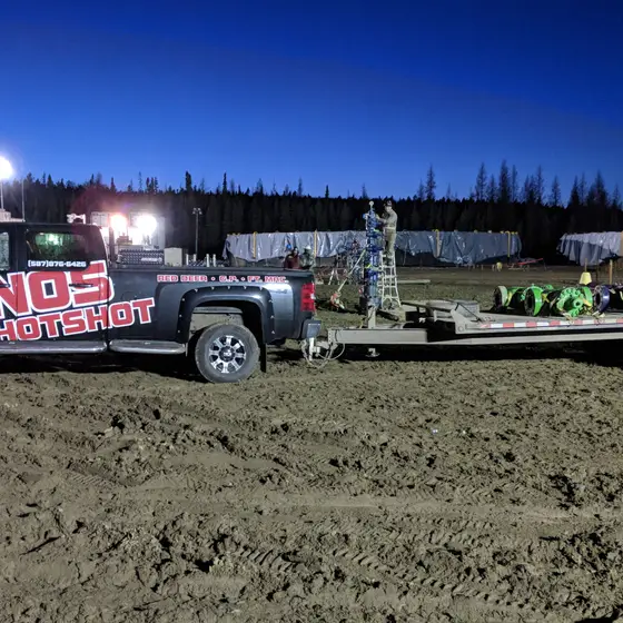 NOS Hotshot truck hooked to a loaded three-axle gooseneck deck on a muddy lease site at dusk, light towers and camp trailers behind