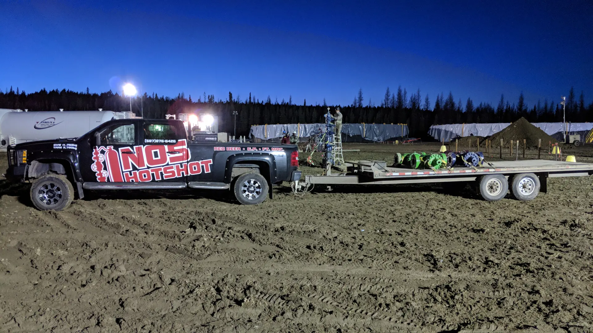 NOS Hotshot truck hooked to a loaded three-axle gooseneck deck on a muddy lease site at dusk, light towers and camp trailers behind
